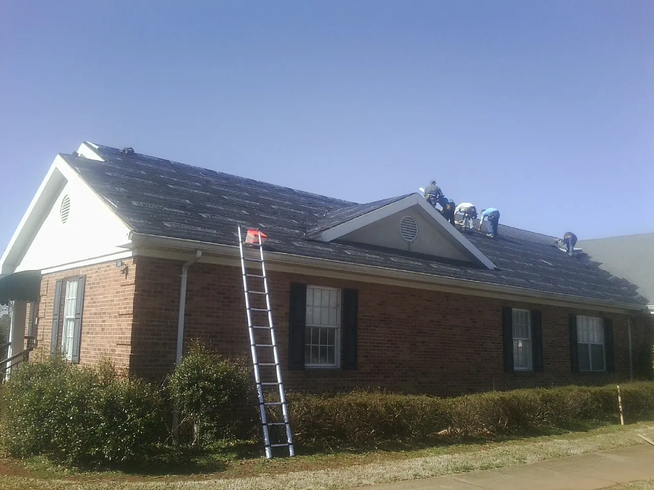Crew tearing shingles off a brick ranch roof with a ladder set — Metal Roof Repair work by our crew in Aliceville