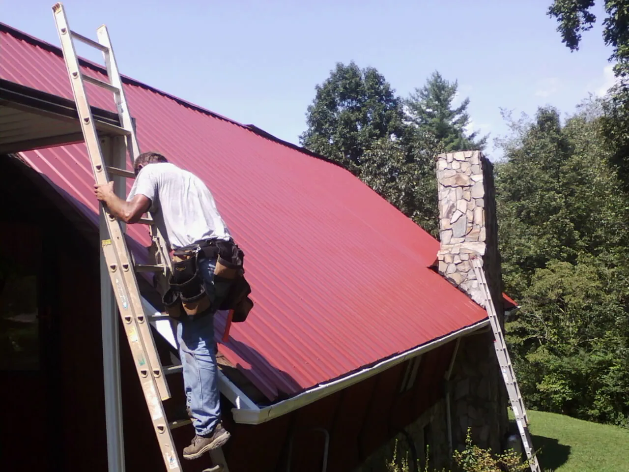 Roofer on a ladder beside a red metal roof and stone chimney — Soffit & Fascia Repair by Roofing Brothers in Aliceville